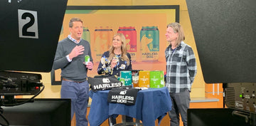 This is a photograph of Paul Pirner, Megan Newquist, and Christ Egert on the television set of KSTP Minnesota Live show. Cartons of Hairless Dog brews and Hairless Dog apparel are on a table in front. 